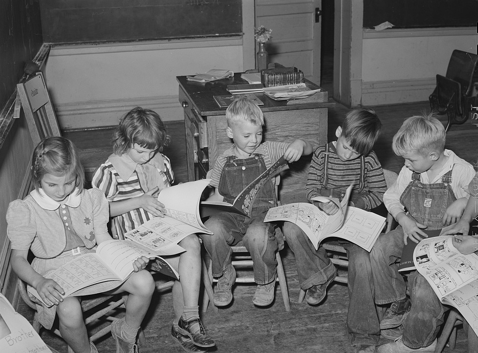 A black-and-white photograph of six schoolchildren sitting close together on small chairs, each bent over an open picture book on their lap