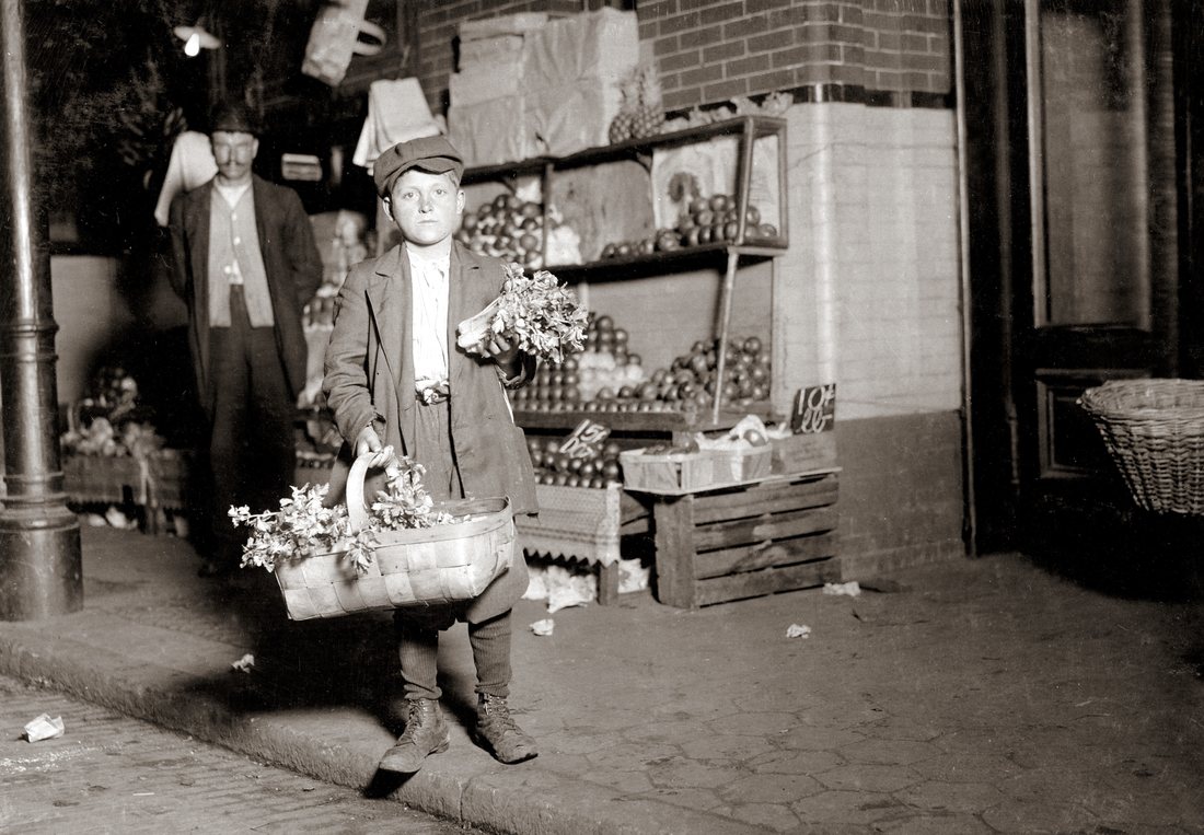 A boy in a cap and coat standing at a market stall at night with bunches of celery, photographed under electric light