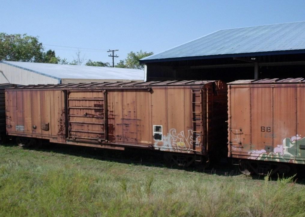 A long enclosed railroad freight car standing on tracks, with a wide sliding door in the middle of its side and a walkway along the roof