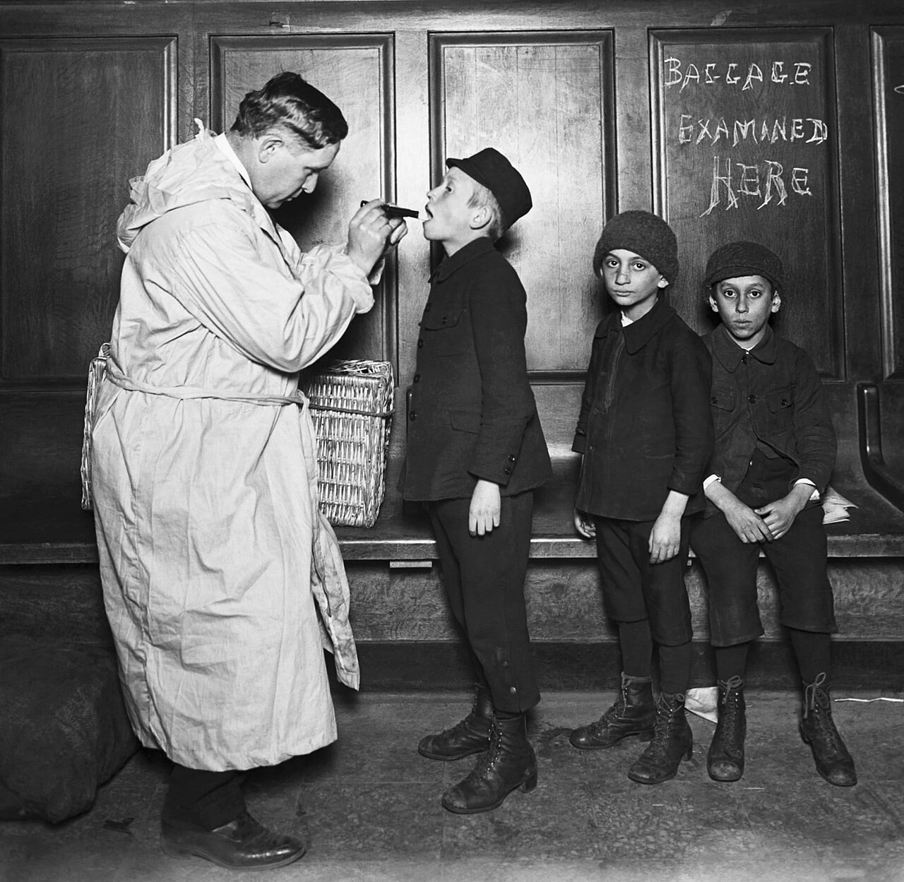 A group of children standing outdoors in coats and hats while a man in a hat leans down to look closely at one child's face