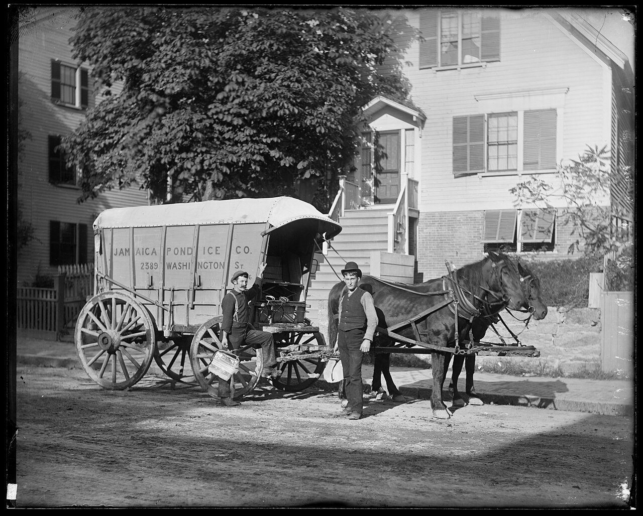 Two men beside a horse-drawn ice wagon on a city street, one lifting a large block of ice with metal tongs onto his shoulder