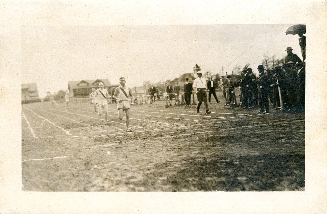 An old photograph of a cinder running track: three runners strung out along the lanes with the leader well clear, an official walking beside them, and a line of spectators in hats pressed against the rail