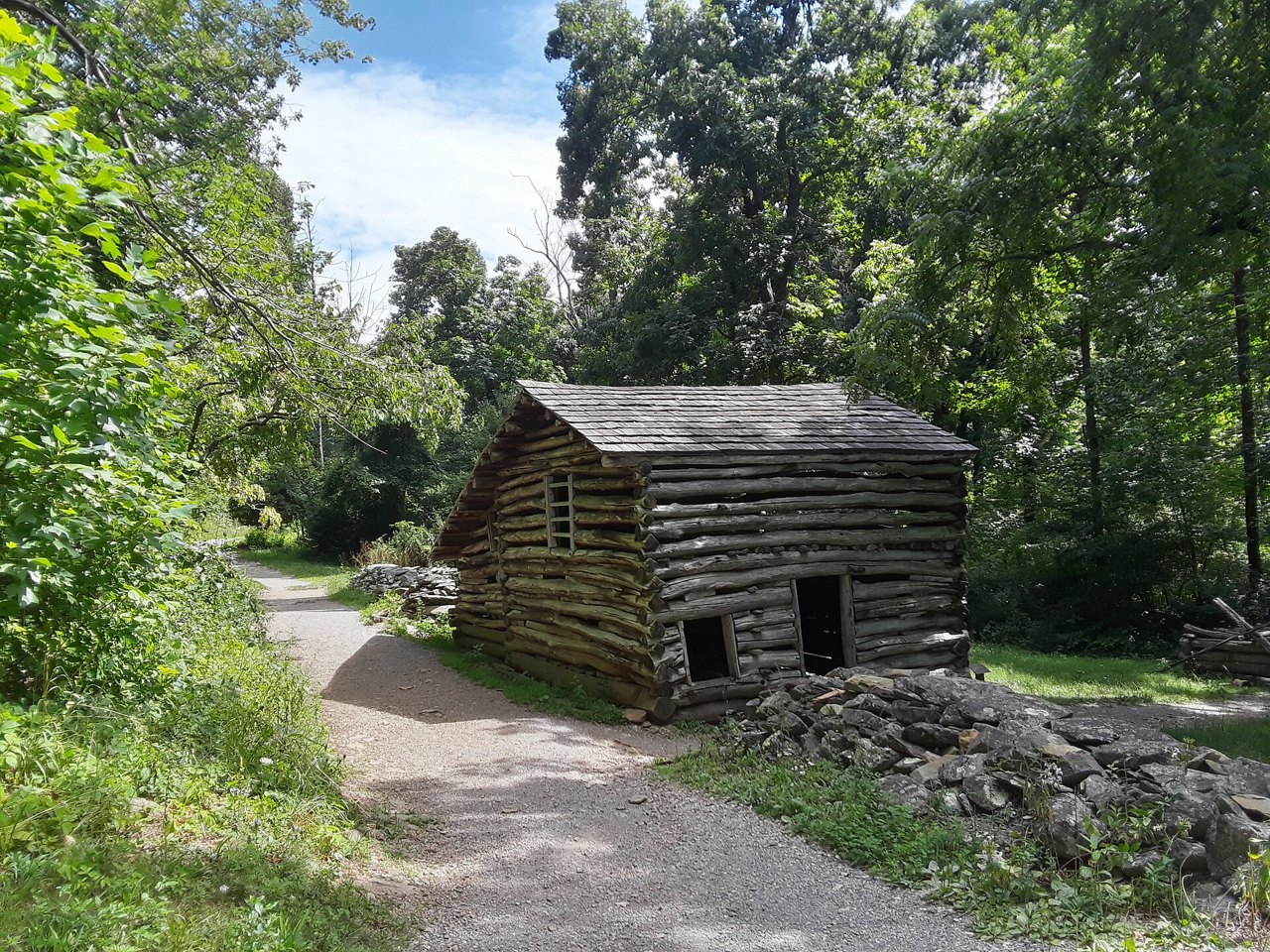 A small, weathered log building with a wood-shingle roof standing beside a gravel path in dense green woods, a pile of rough stones at its corner