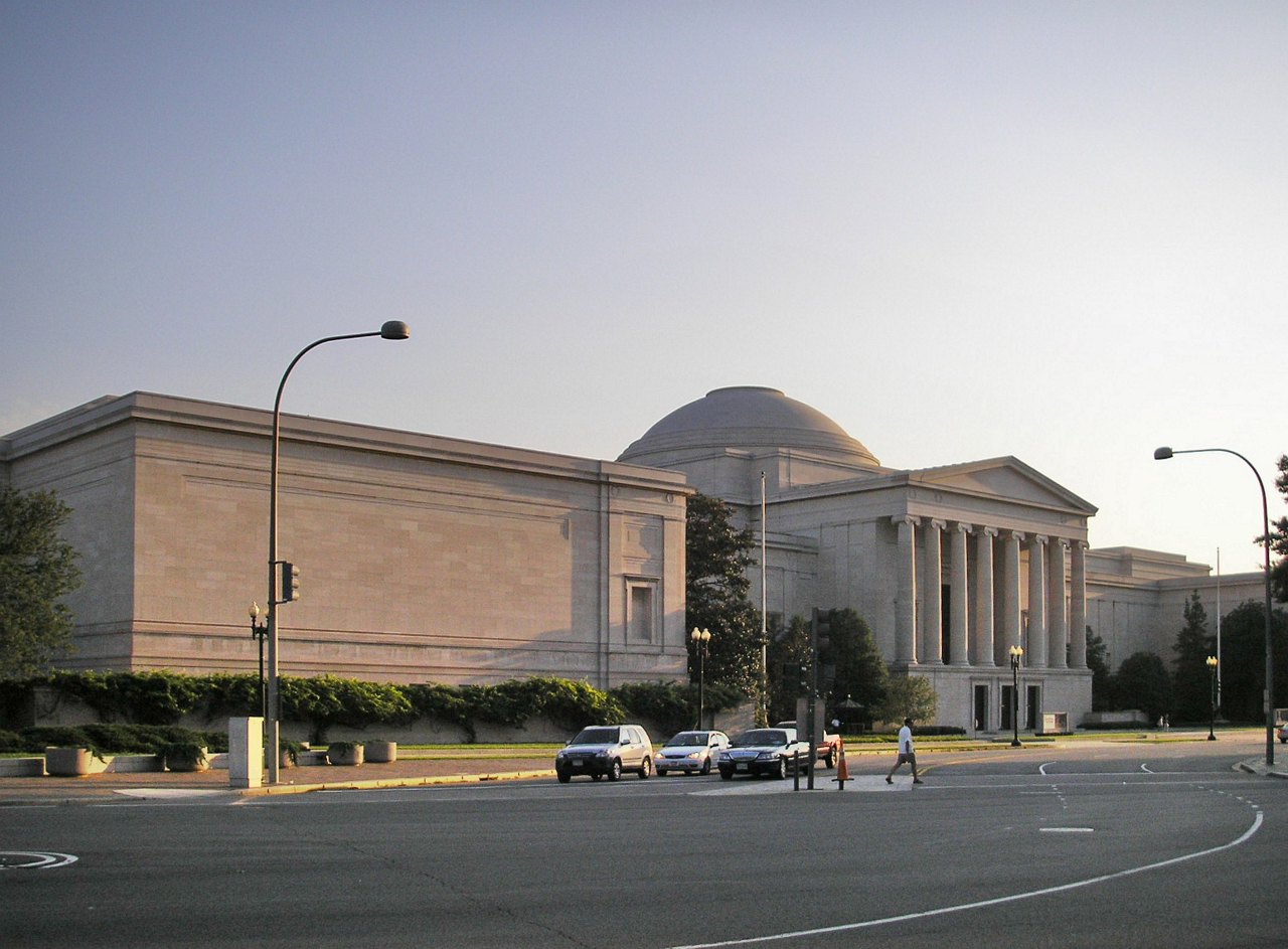 A long, low pale stone museum building with a central dome and a columned entrance, seen across a wide empty road in evening light with a few parked cars