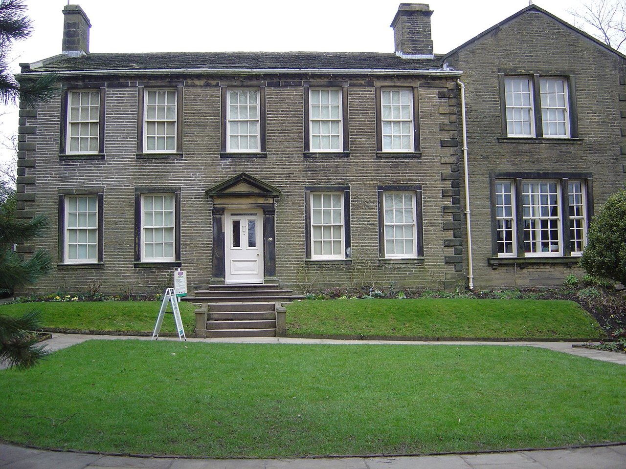 A plain two-story stone house with many sash windows, a small stone porch over the front door, and a neat lawn in front, photographed under a gray sky
