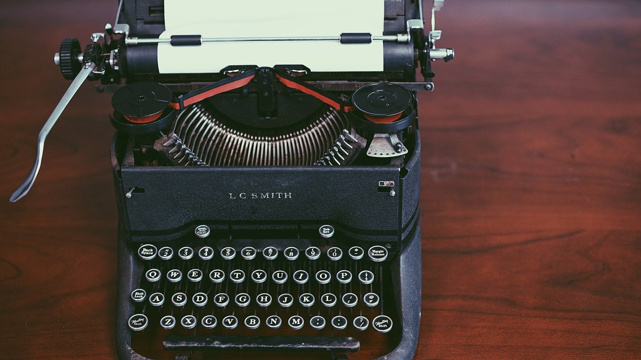 An old black L C Smith typewriter with round glass keys and a sheet of paper in the roller, standing on a dark wooden table
