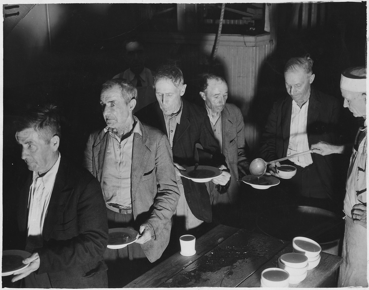 A line of men in worn jackets holding shallow bowls as soup is ladled from a pot at a long wooden table
