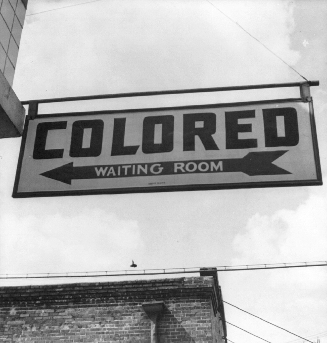 A large painted sign hanging above a street, reading COLORED WAITING ROOM with an arrow pointing left