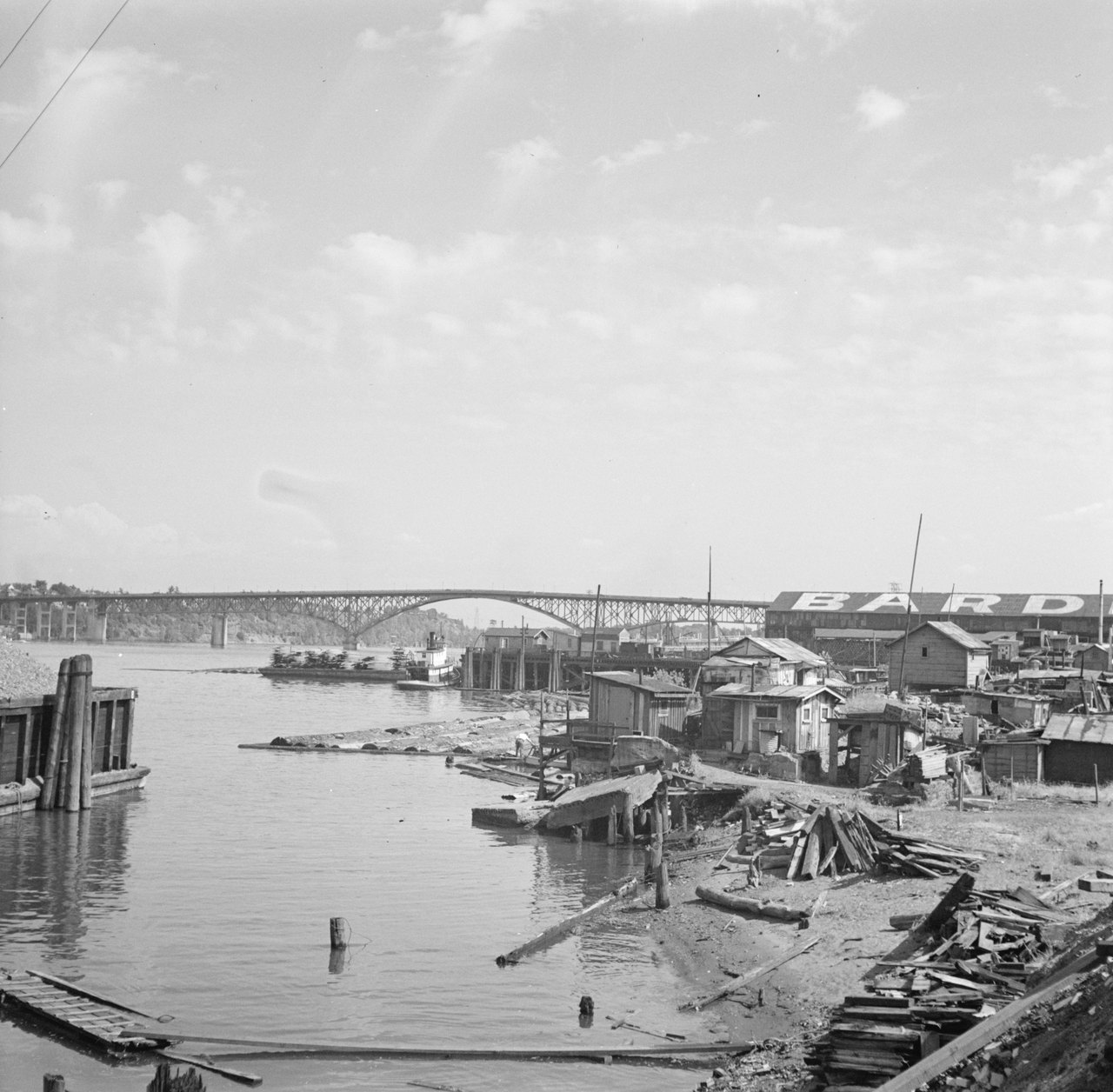 A row of shacks built from scrap boards and tin along a riverbank, with a steel bridge and warehouses behind them