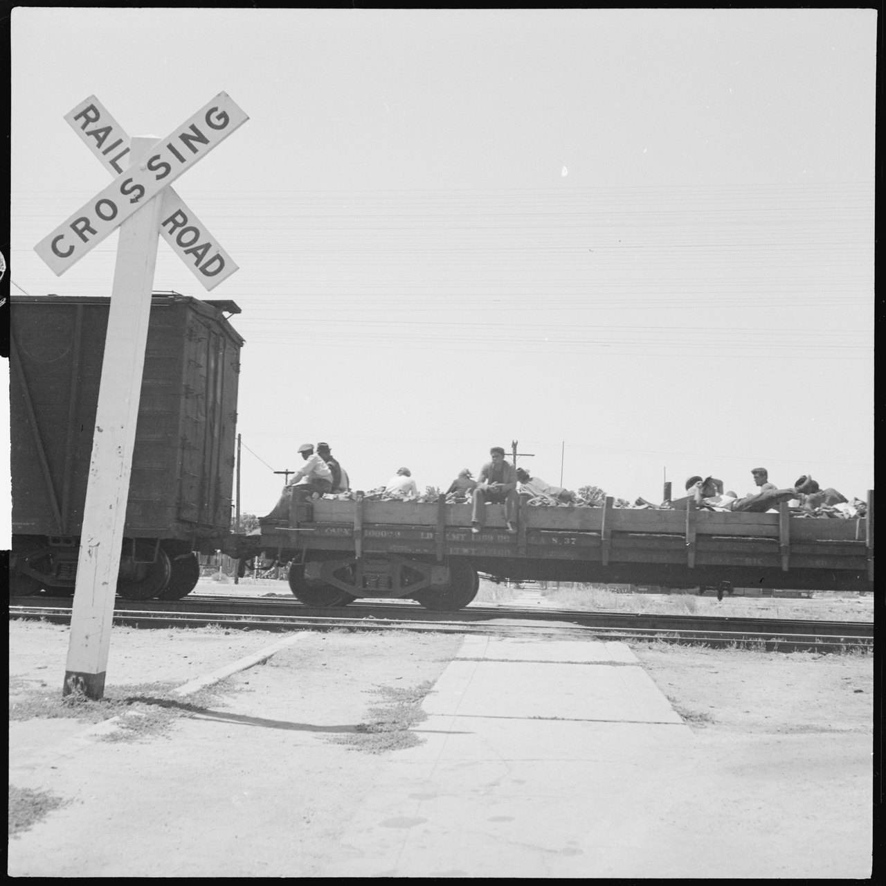 A freight train passing a railroad crossing, with a dozen men sitting and lying on the open flat car behind the boxcars
