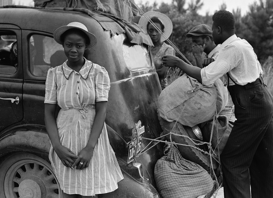 A family beside a loaded car on a roadside, bundles tied with rope to the back, a young woman in a straw hat standing in front
