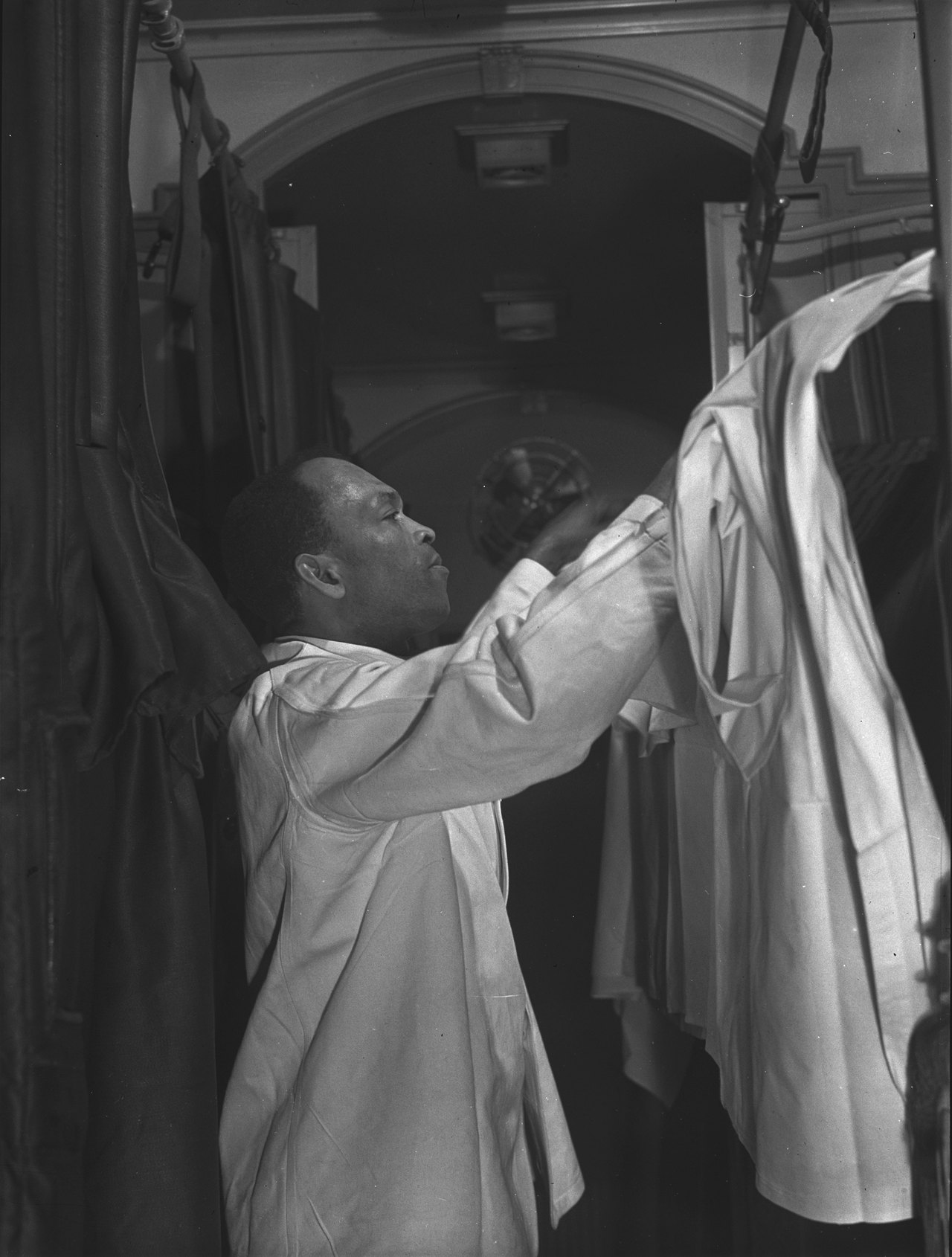 A porter in a white jacket reaching up to fold sheets into an upper sleeping berth in a narrow railroad car aisle