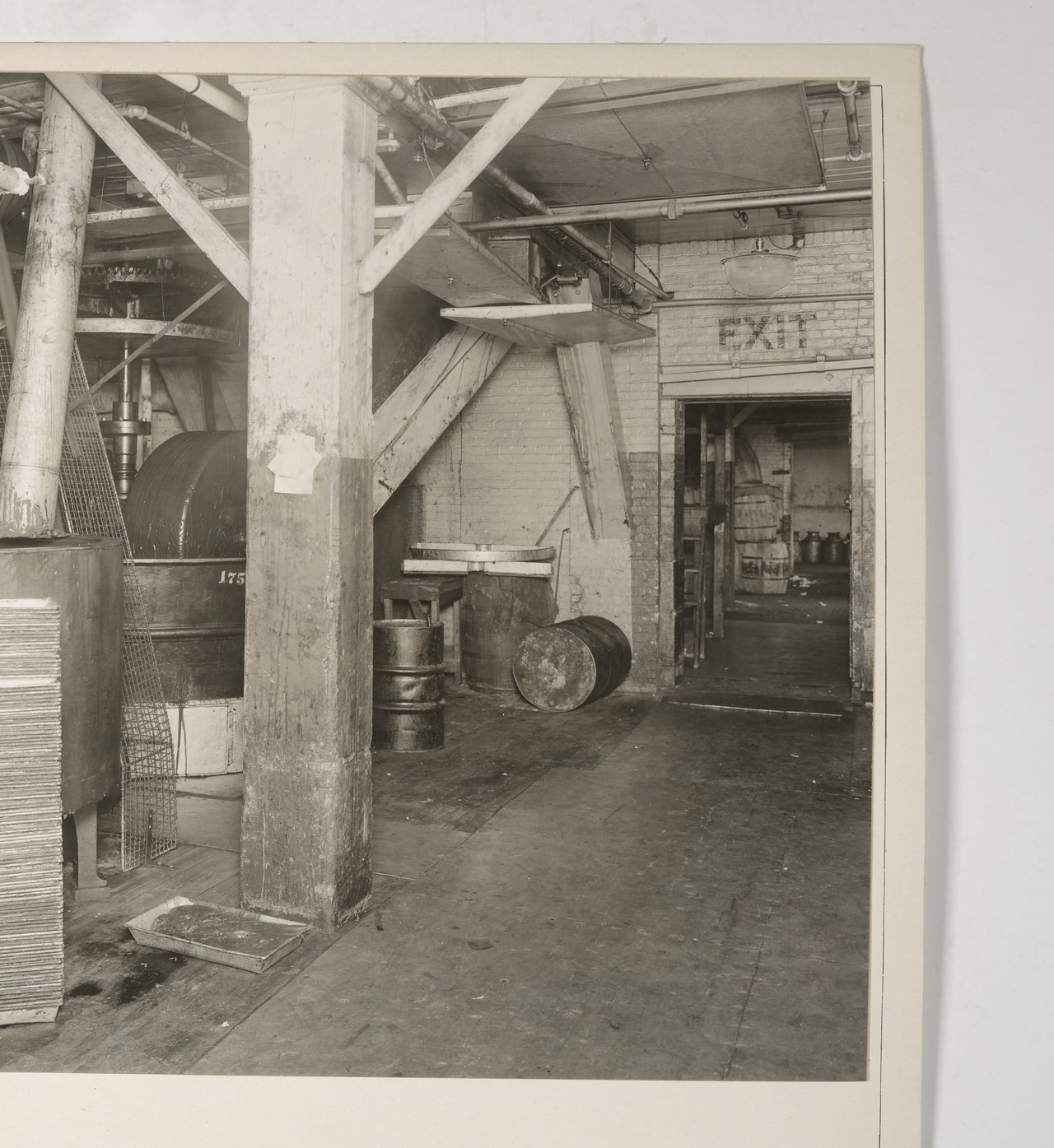 The interior of an early twentieth-century chocolate works, with a large covered mixing drum, stacked metal trays, chutes overhead and an open doorway marked exit