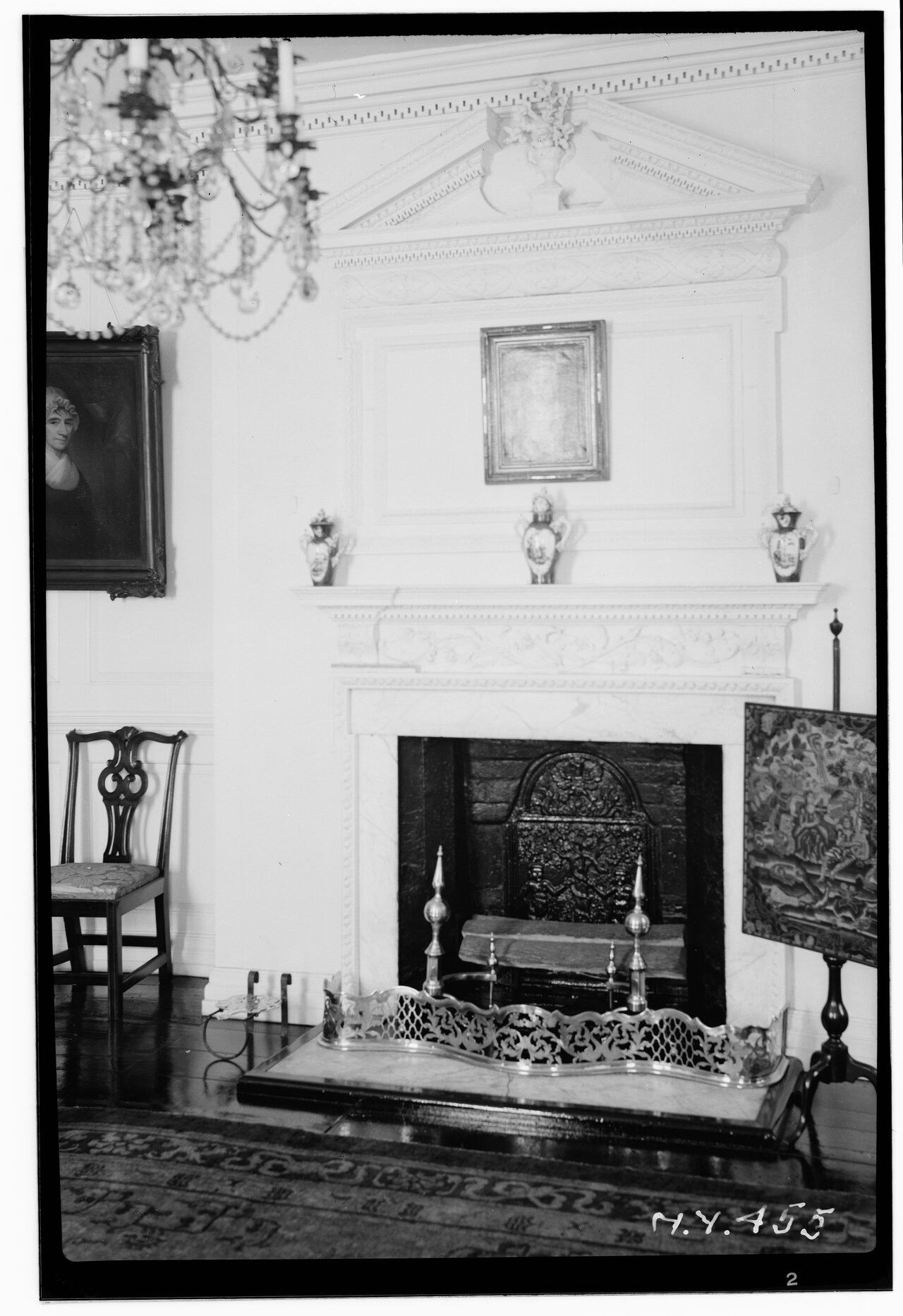 A black-and-white photograph of a paneled parlor: a marble mantel with a carved pediment above it, a cut-glass chandelier, porcelain jars on the shelf, a portrait in a heavy frame, an armchair and a brass fender at the hearth