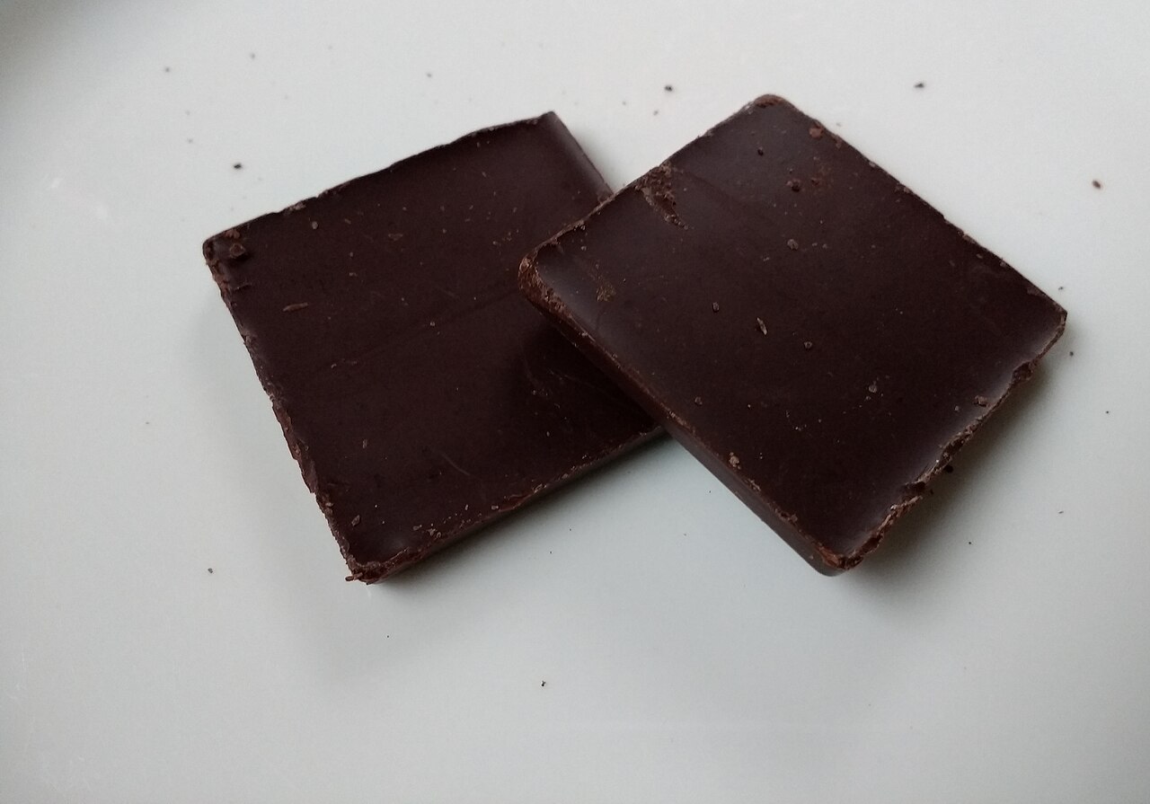 Two thick squares of dark chocolate lying on a plain pale surface, one overlapping the other, their broken edges rough where they were snapped apart