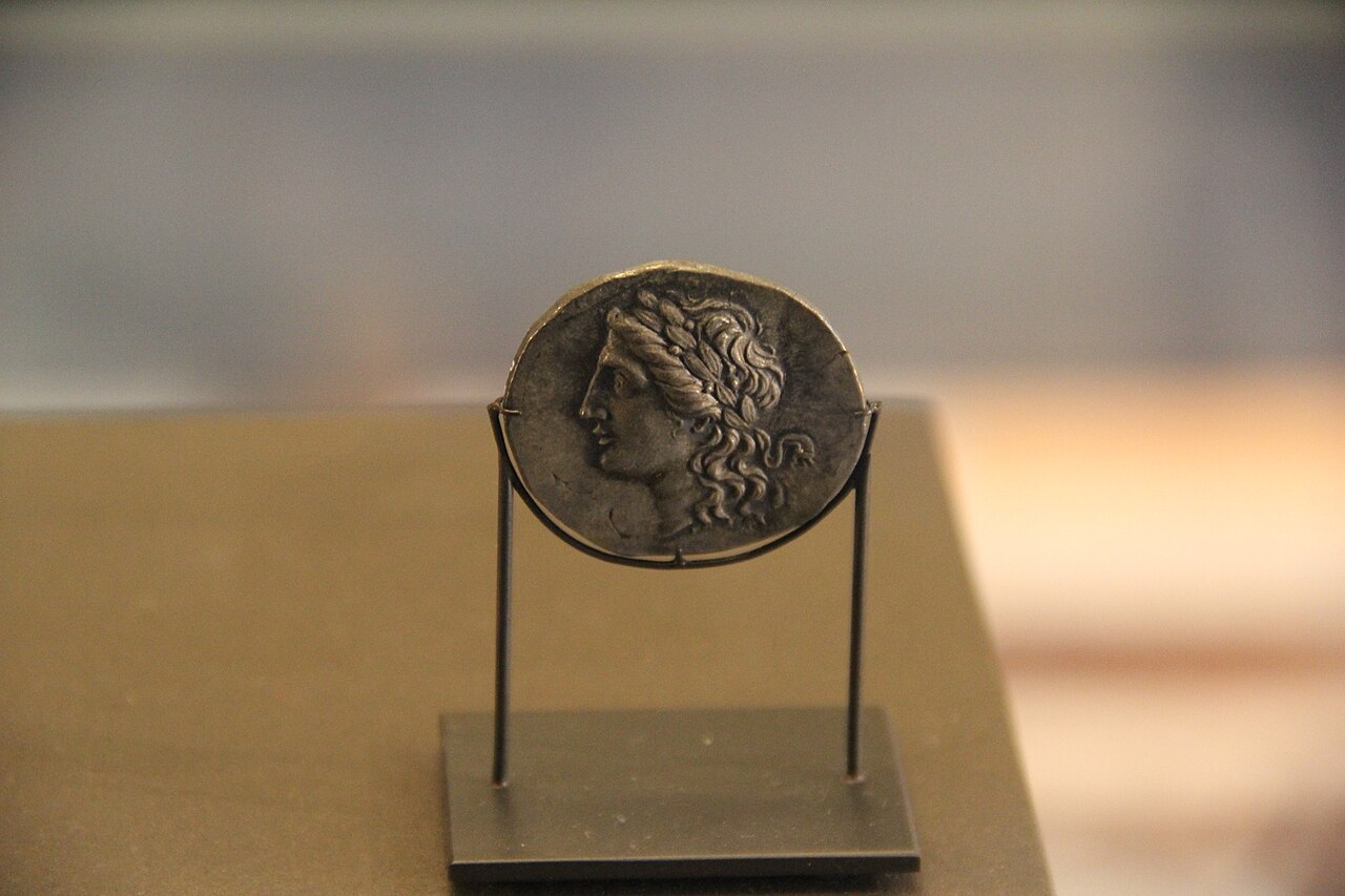 An ancient silver coin lying on a dark surface, worn smooth at the edges, with a stamped head in profile still visible on its face