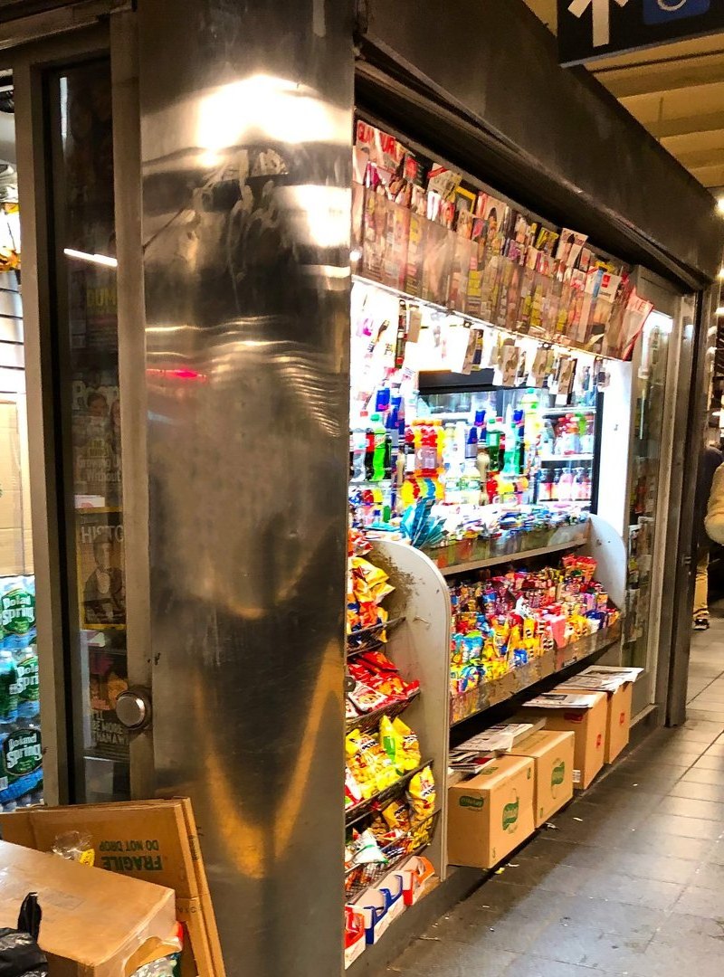 A narrow steel-and-glass newsstand booth inside a subway station, its front packed with magazines, candy, snacks and bottled drinks beside a tiled pillar
