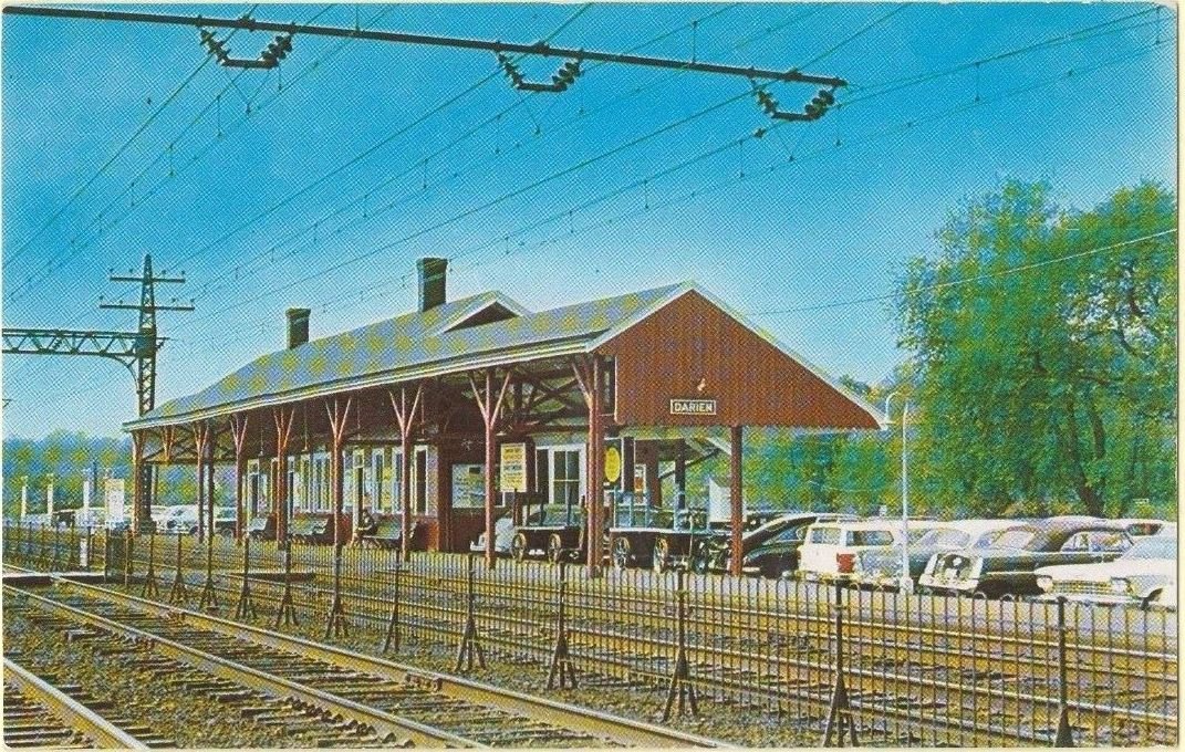 A colored midcentury postcard of a small brown railroad station beside several tracks, with overhead electric wires on tall poles and cars parked beyond the platform