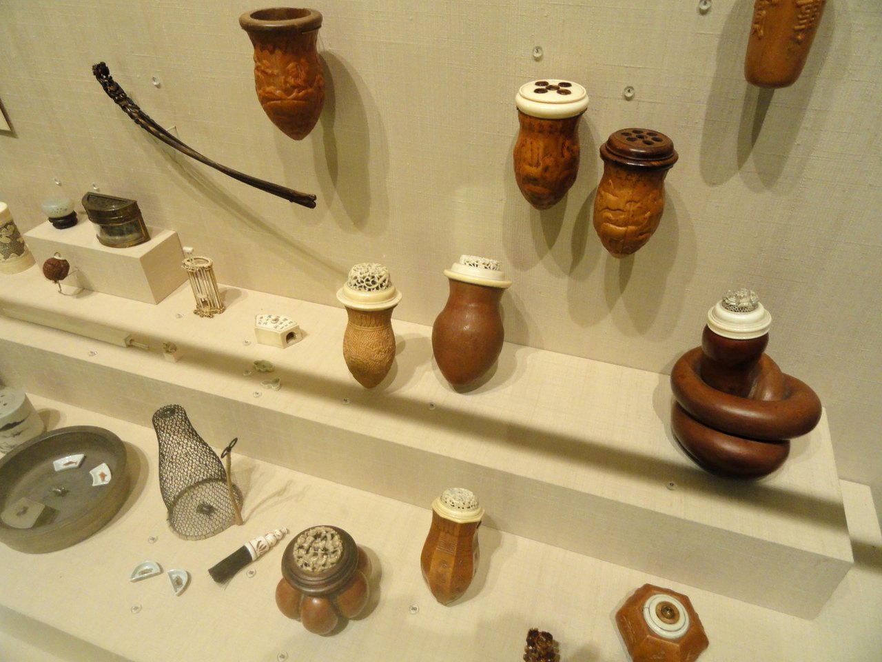 A museum wall case holding carved brown gourds with pierced lids, small round dishes, a tiny wire cage and slender tools, all used for keeping crickets
