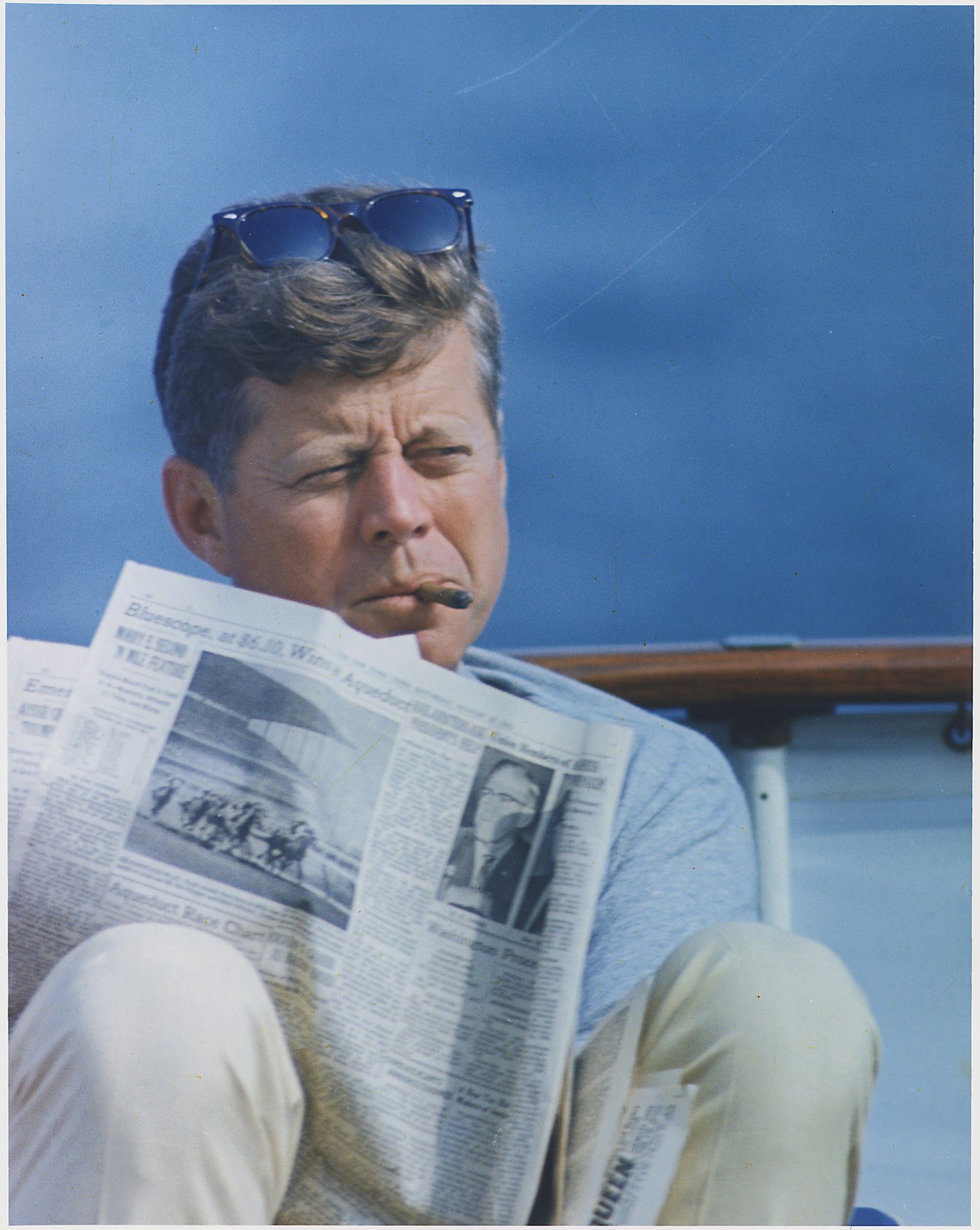 A man in a gray shirt with sunglasses pushed up on his hair sits on a boat deck against blue sky and water, holding an open copy of the New York Times