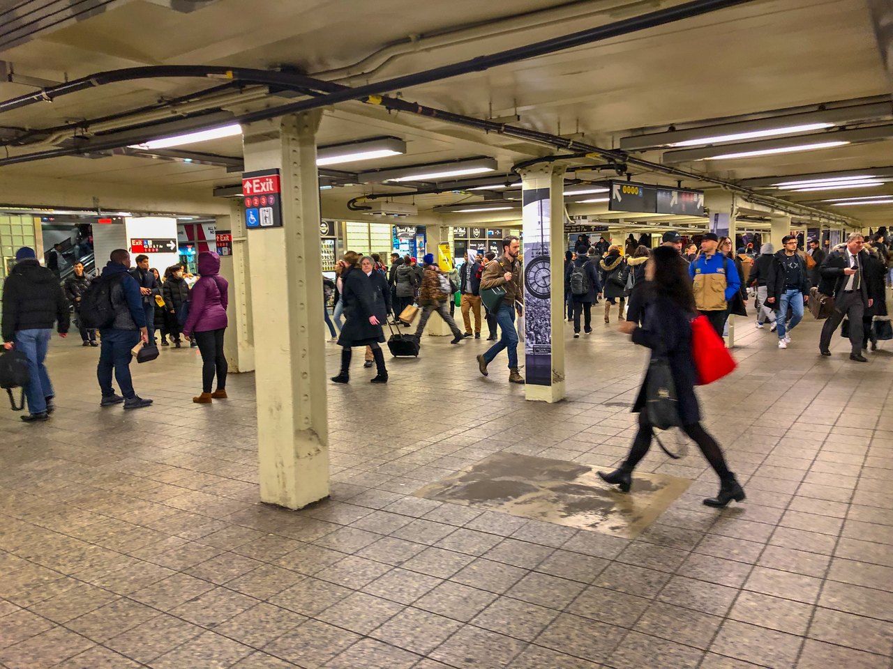 A wide, low-ceilinged subway mezzanine with a tiled floor and pillars, crowded with people in winter coats walking in every direction under fluorescent lights