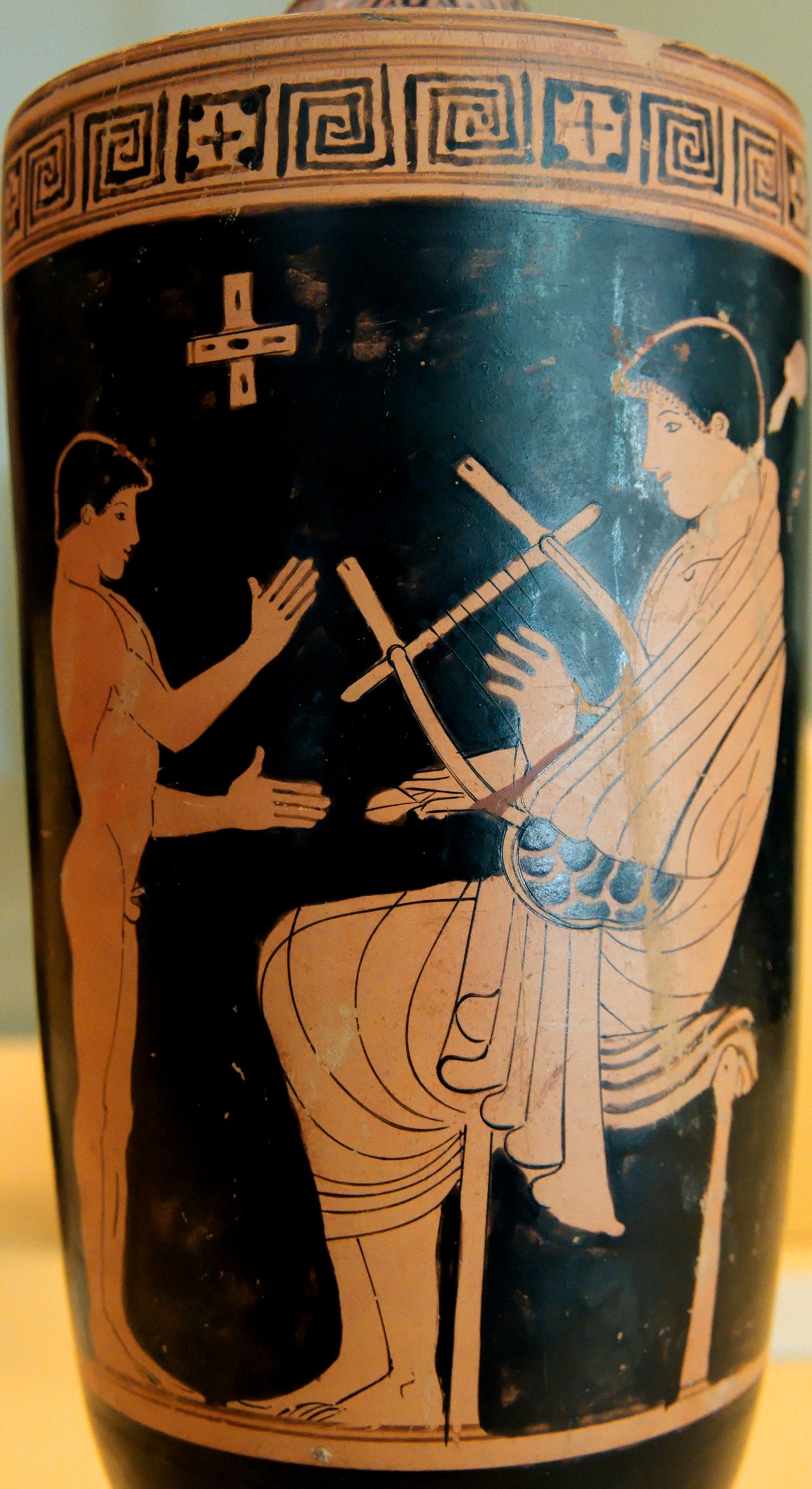 A red-figure Greek vase painting of a seated figure holding a small stringed lyre in the lap while a standing figure raises a hand toward it