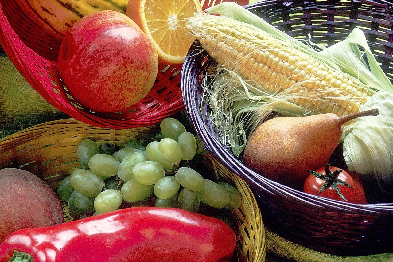 Three woven baskets seen close up, holding green grapes, a peach, a halved orange, an ear of corn with the husk pulled back, a pear, a tomato and a large red pepper
