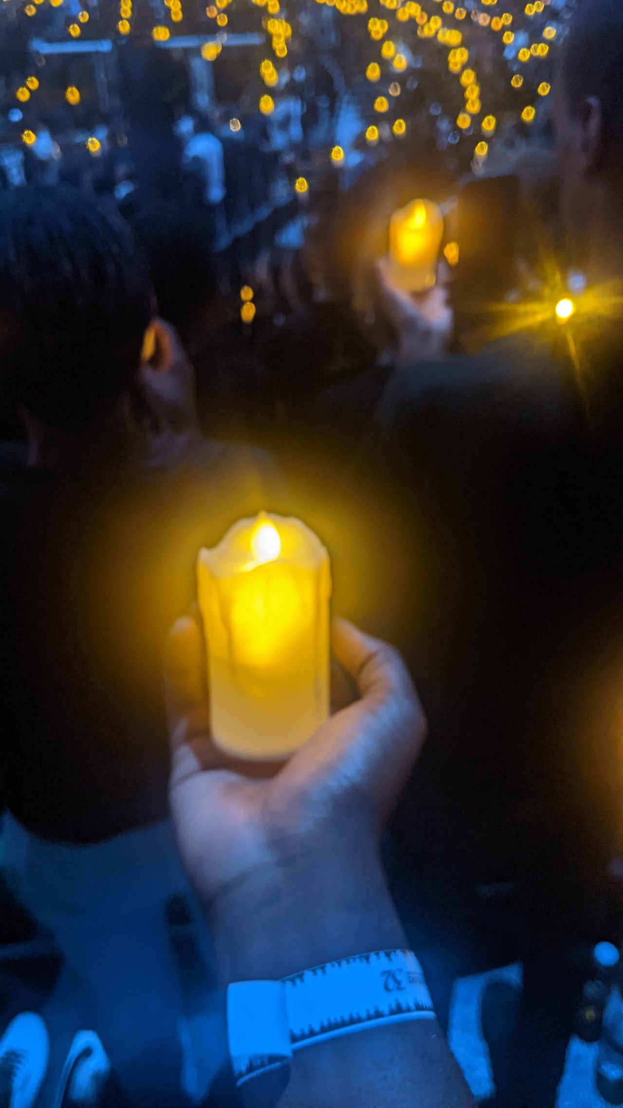 A hand held up in a dark crowd cupping a single lit candle, with other small points of flame glowing faintly among shadowed figures behind