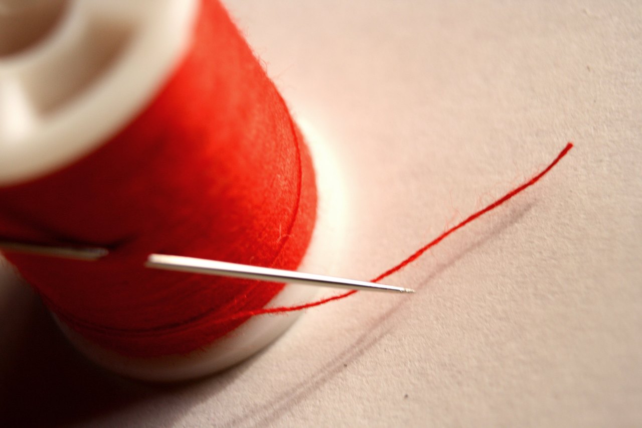 A close view of a steel sewing needle lying on pale cloth beside a spool of bright red thread, a single strand trailing away from the eye