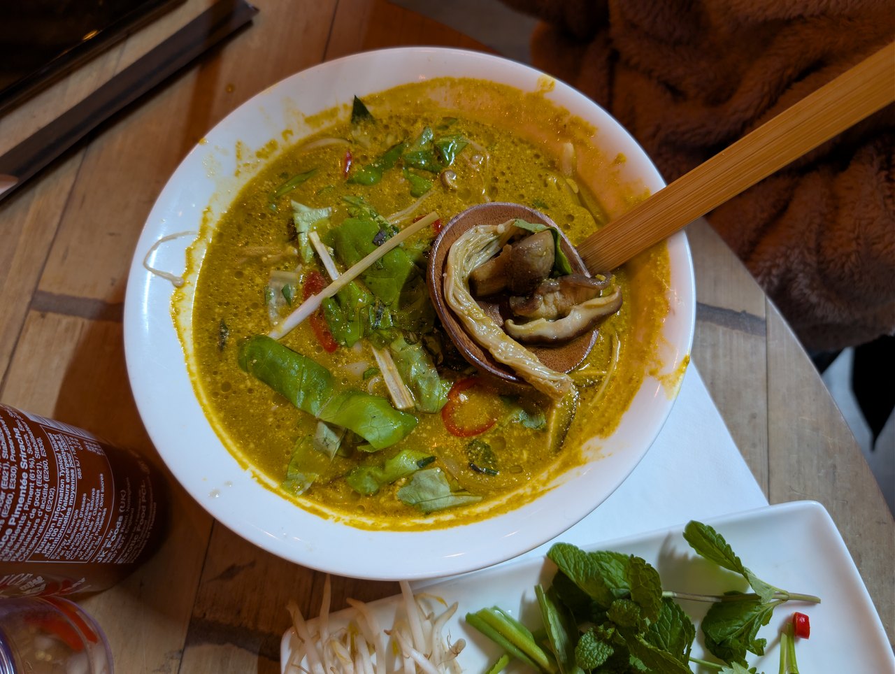 A wide white bowl of steaming golden noodle soup seen from above, crowded with mushrooms, green herbs and sliced chili, with chopsticks resting across the rim
