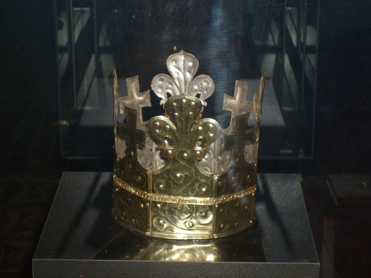A tall gold crown with pointed decorated bands standing alone on a dark plinth inside a lit glass museum case
