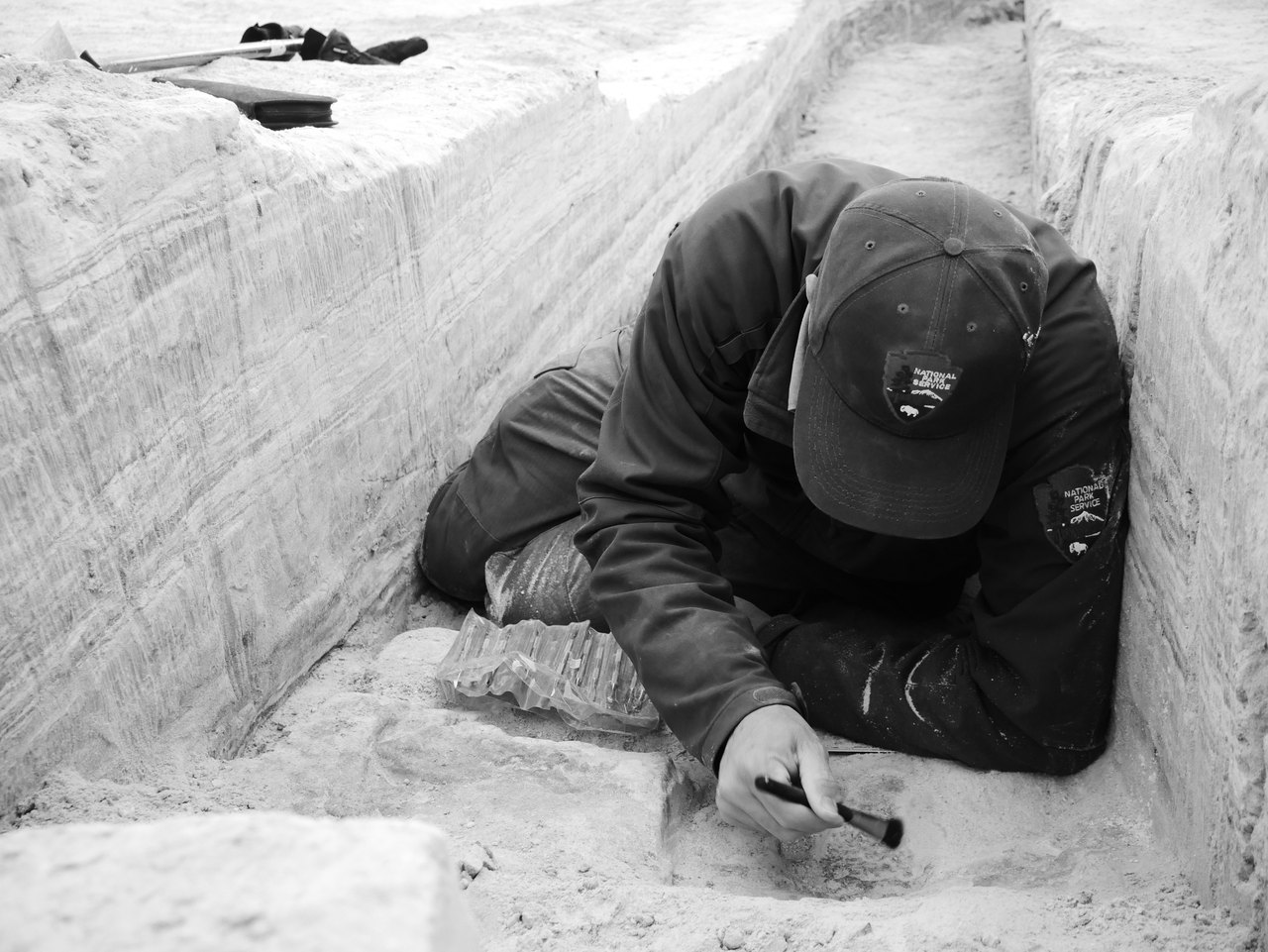 A researcher lies along the floor of a narrow excavation trench, brushing carefully at the pale sediment of a stepped wall beside them