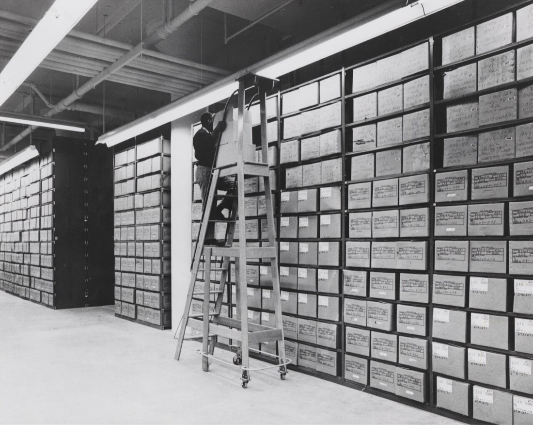 A black-and-white photograph of a records center: walls of steel shelving packed with labeled cartons rise floor to ceiling, and a worker on a tall rolling ladder reaches for one box