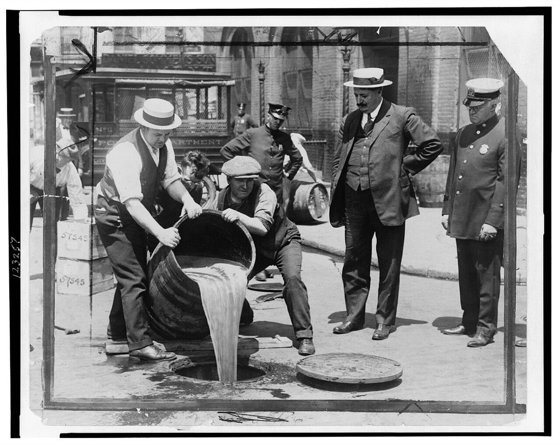 A black-and-white photograph of men on a city street tipping a large barrel so its contents pour into an open manhole, while a police officer and a heavyset official in a straw hat stand watching