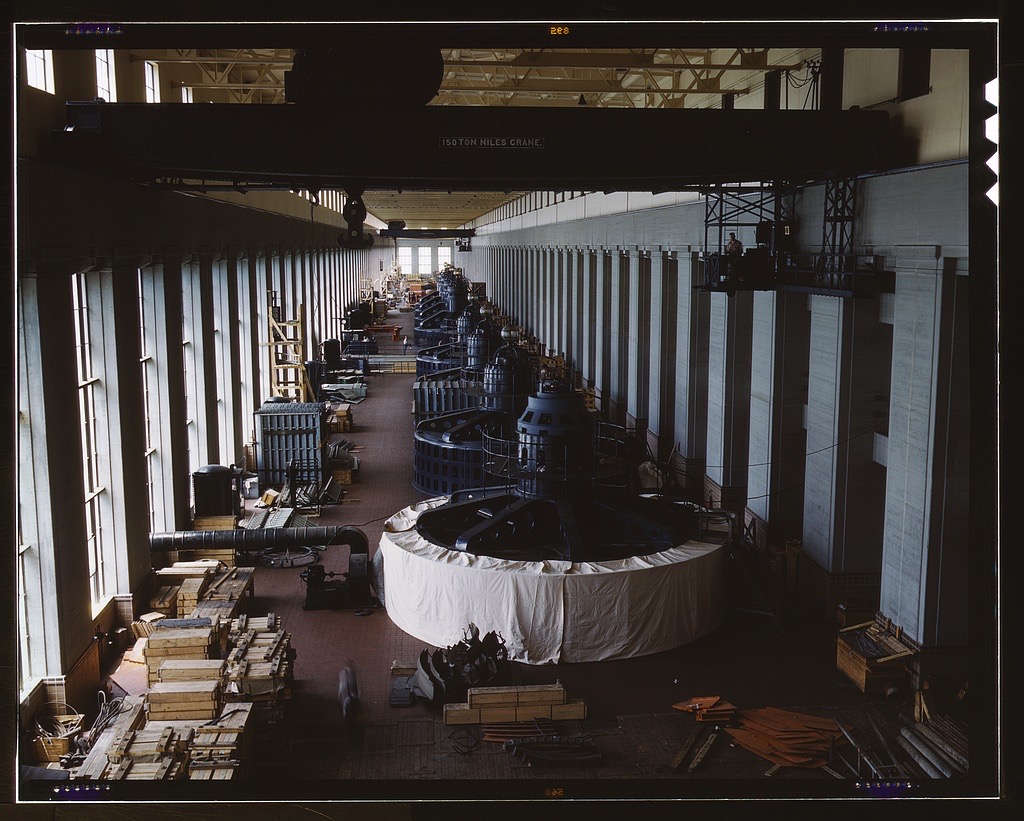 A vast powerhouse hall photographed in color: a row of dark round generator units recedes down the room, the nearest one wrapped in a white cover, with wooden crates on the floor and a crane bridge overhead