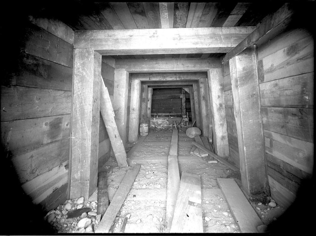 A black-and-white photograph looking down a mine tunnel framed by heavy squared timbers, one set behind another, with planks on the rocky floor and a barrel and wheelbarrow at the far end