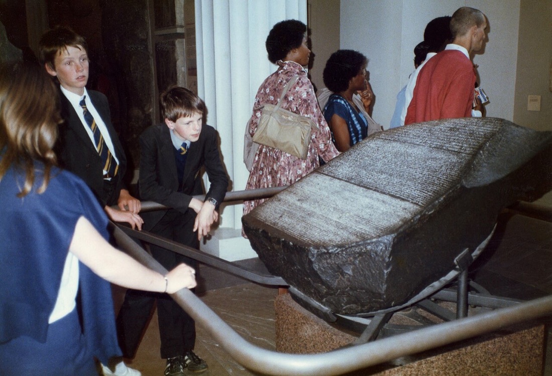 A color photograph from 1985 of the Rosetta Stone lying tilted on a display cradle in the British Museum, its dark surface covered in bands of close-cut inscription, with visitors and two schoolboys standing around the railing