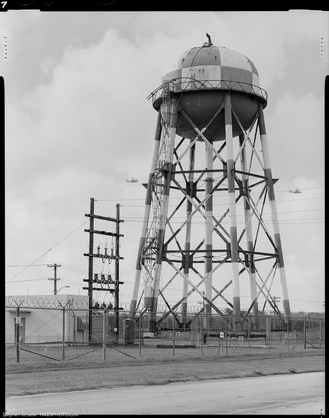 A black-and-white photograph of a tall elevated water tank: a huge sphere painted in a checkerboard pattern standing on braced steel legs, with power poles and a chain-link fence below