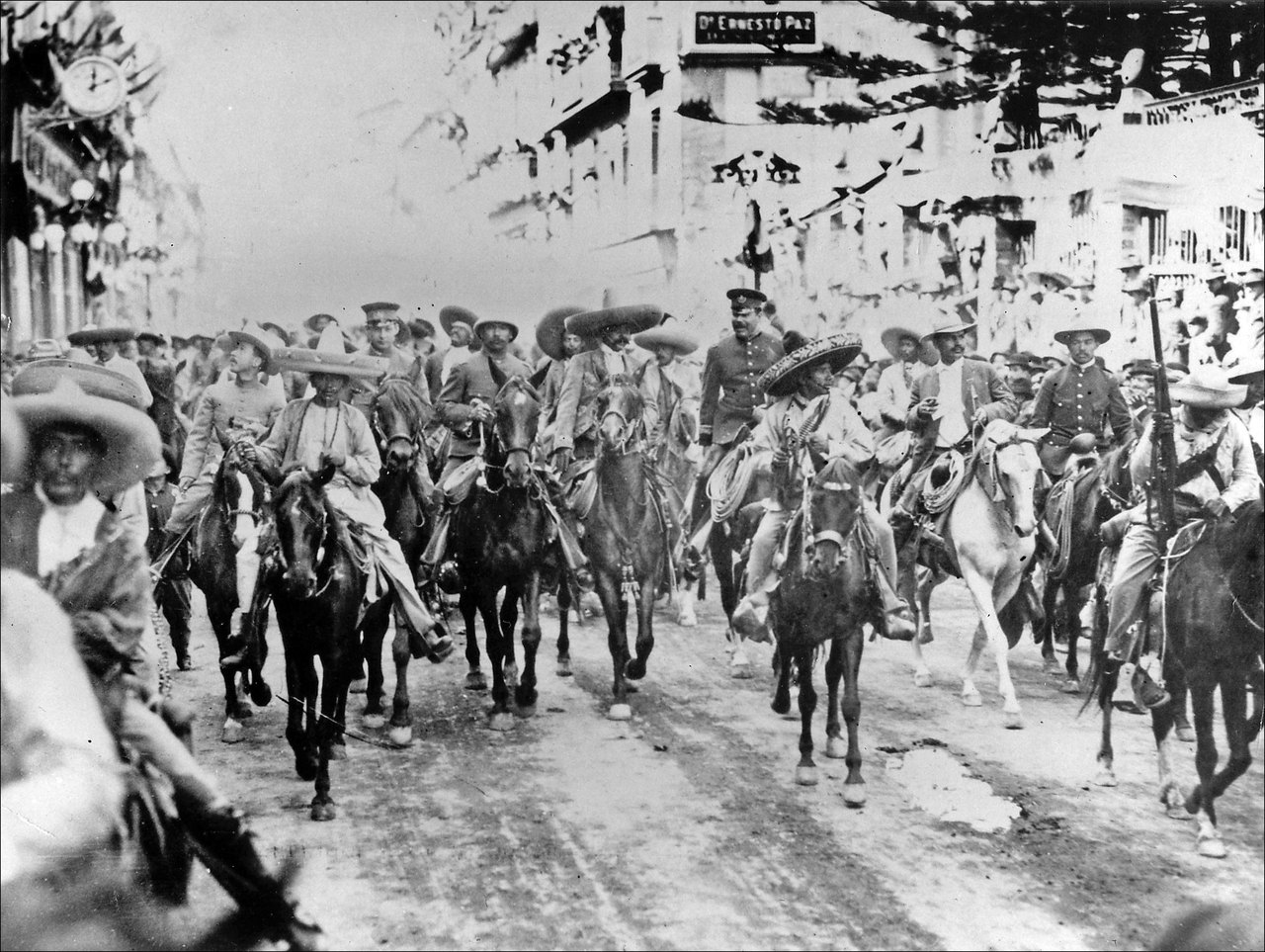 A long column of mounted revolutionary fighters in wide-brimmed hats riding down a decorated town street, watched by crowds on the balconies