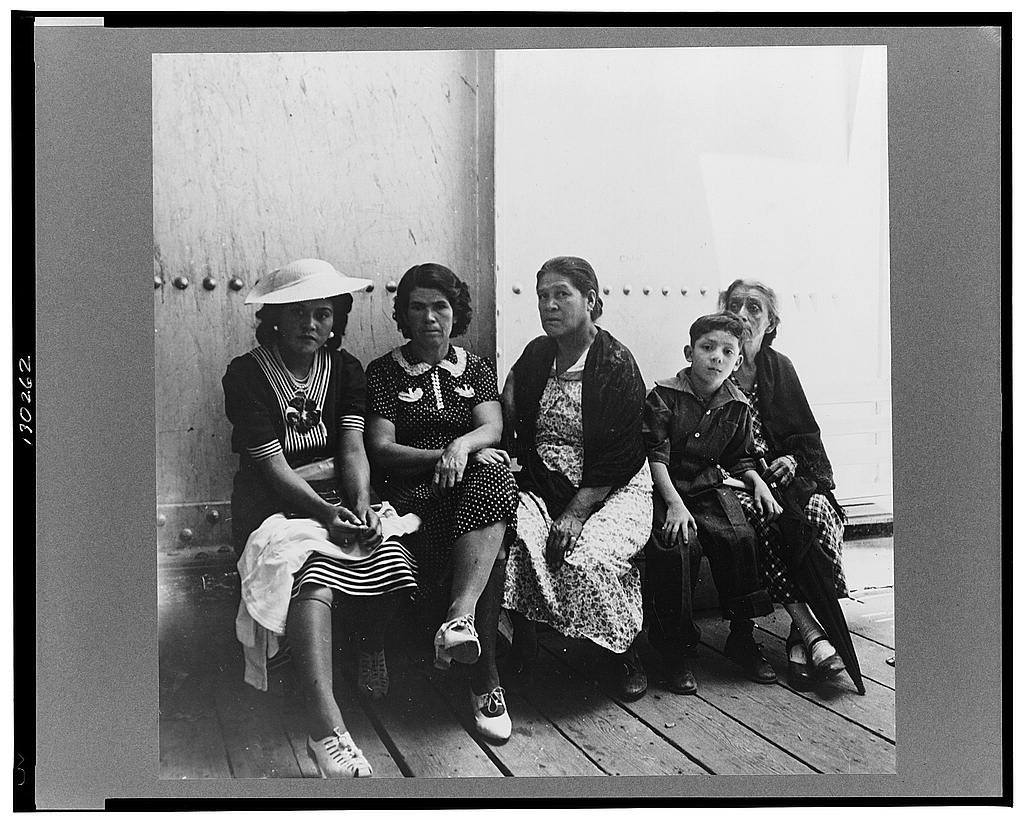 Four women and a boy sitting close together on a bench against a plank wall at a border inspection station, waiting, their belongings beside them