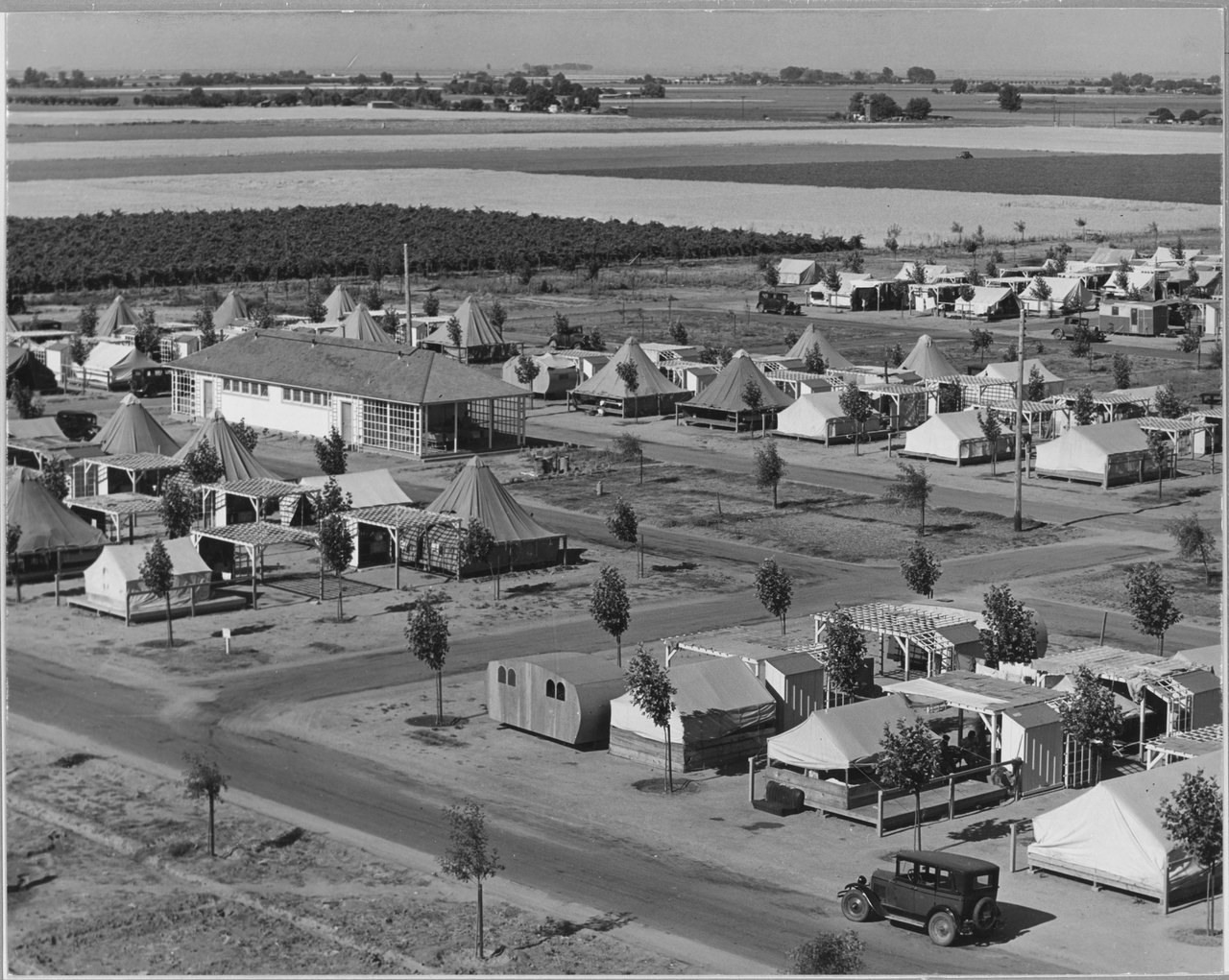 A view from above of a farm labor camp: long straight rows of tent cabins and small wooden shelters on bare ground, with flat farmland stretching to the horizon behind