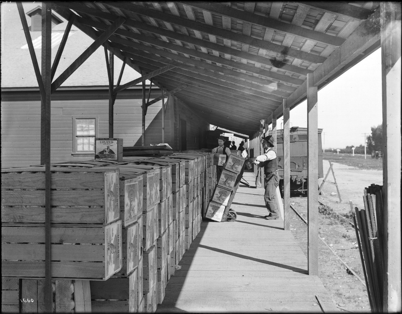 A long covered loading platform stacked with wooden fruit crates, two workers moving crates on a hand truck toward a railroad car waiting alongside