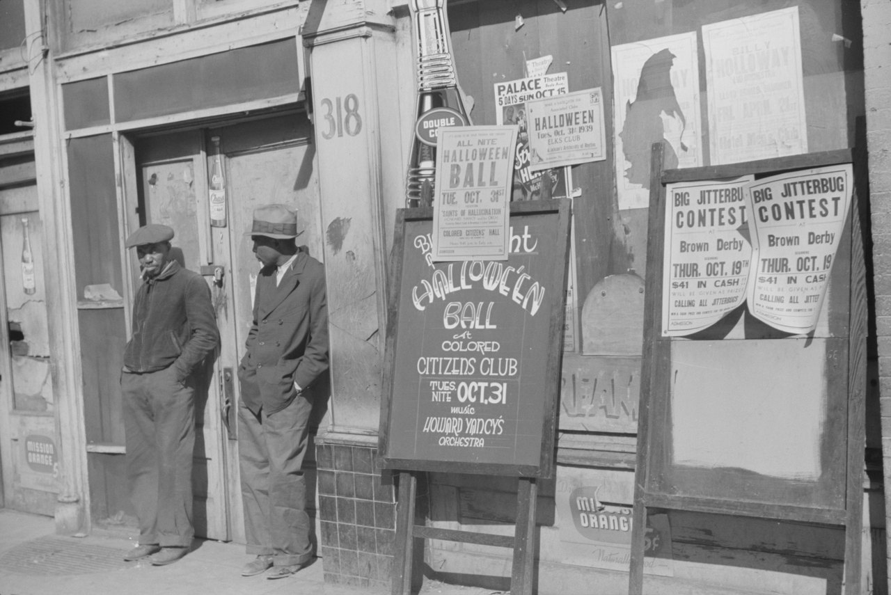 Black-and-white street photograph of two men in caps and jackets standing beside a shop doorway numbered 318, with hand-lettered posters for a Halloween ball and a jitterbug contest propped on the pavement