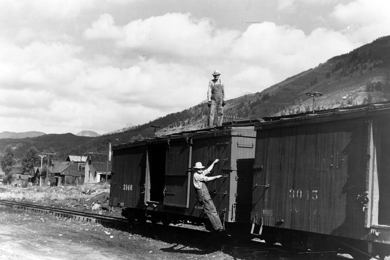 Black-and-white photograph of two wooden freight boxcars on a track below a mountainside; one man in overalls stands on the roof and another climbs the ladder beside an open sliding door