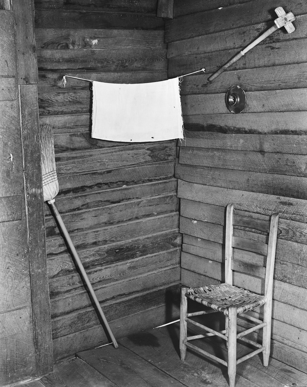 Black-and-white photograph of the bare corner of a wooden cabin: rough plank walls, a single straight chair, a broom leaning in the corner, and a small towel hanging on a line