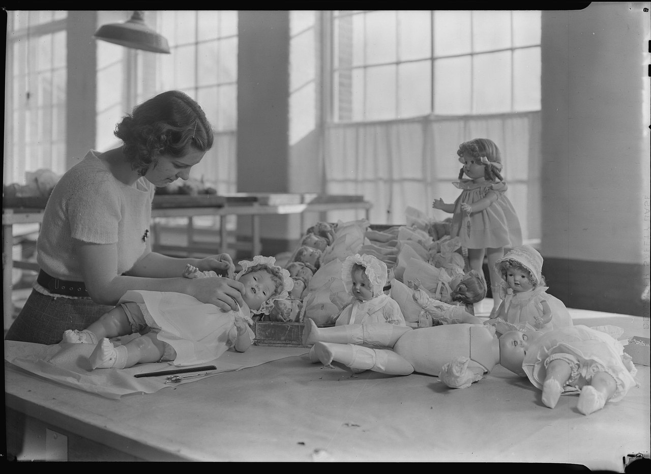 Black-and-white photograph of a young woman at a long worktable dressing baby dolls, with several finished dolls lying and standing around her under tall factory windows