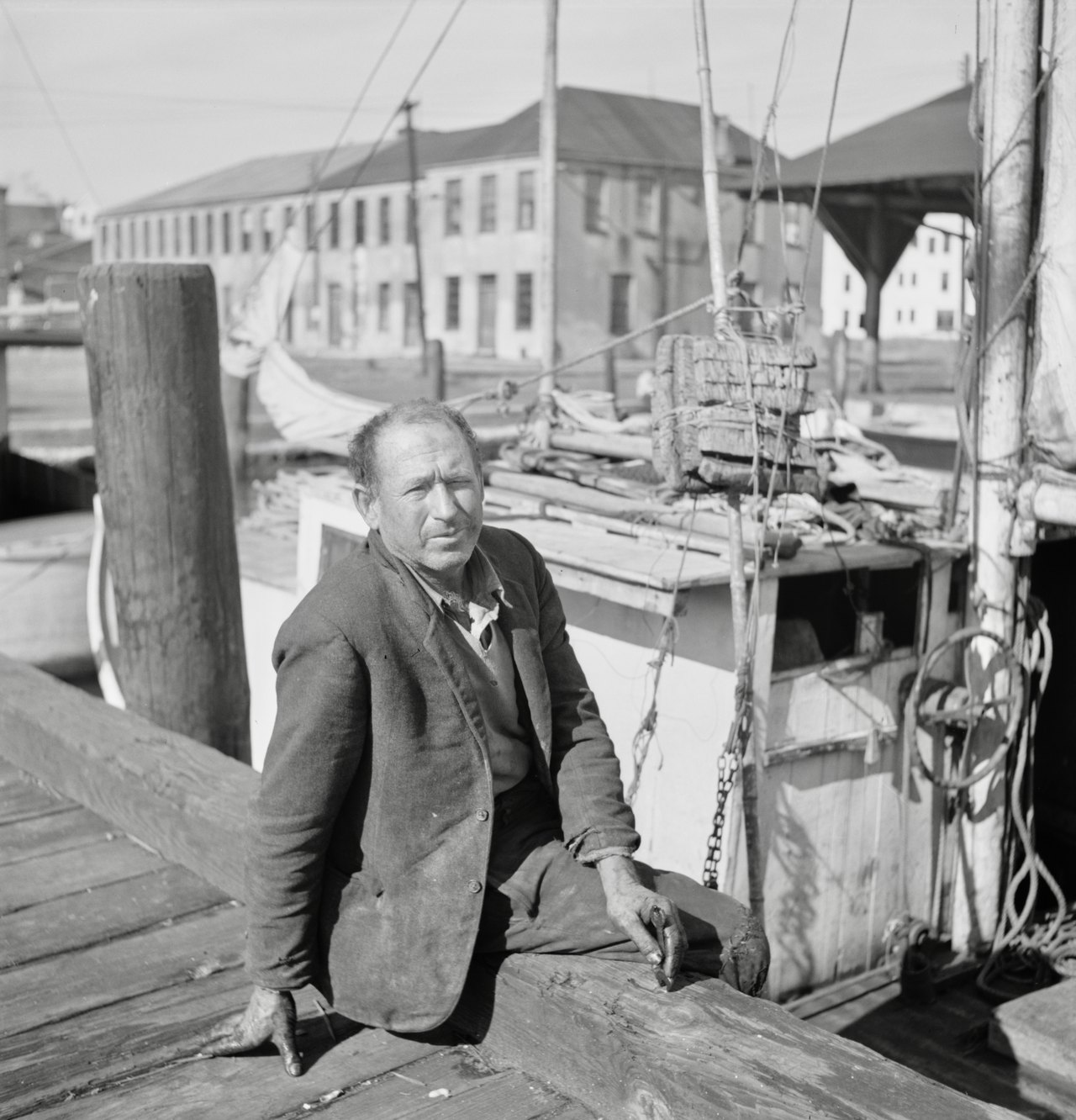 Black-and-white photograph of a weathered man in a dark jacket sitting on a wooden dock beside a fishing boat, with masts, ropes, and a low warehouse behind him