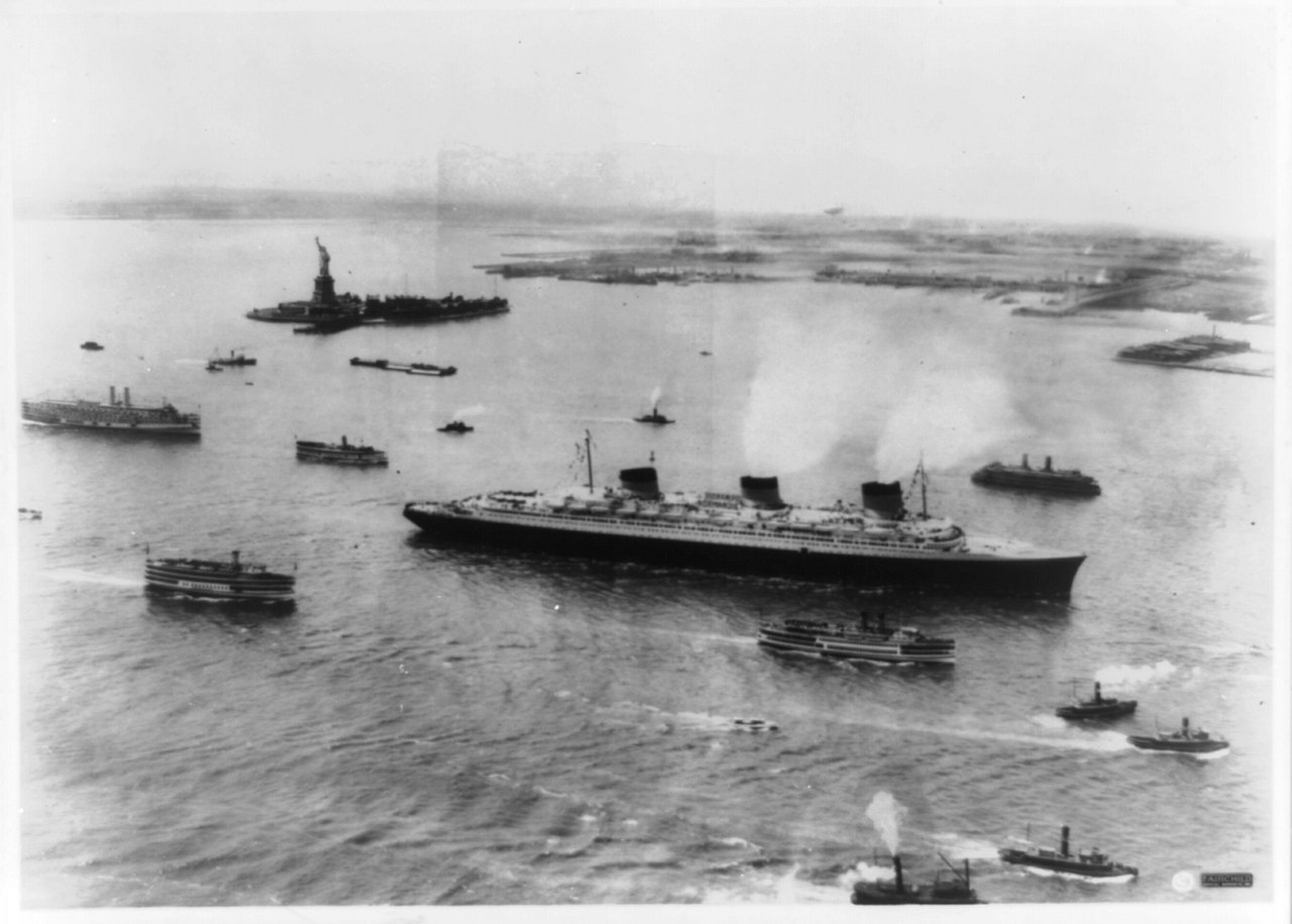Aerial black-and-white photograph of a huge three-funneled ocean liner steaming into a harbor surrounded by small tugboats, with the Statue of Liberty visible on an island at the upper left