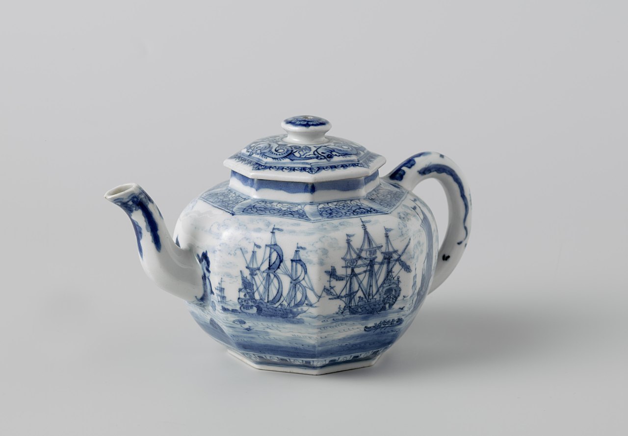 An eight-sided white porcelain teapot with a domed lid, painted in blue with tall European sailing ships on the sea, photographed against a plain gray background