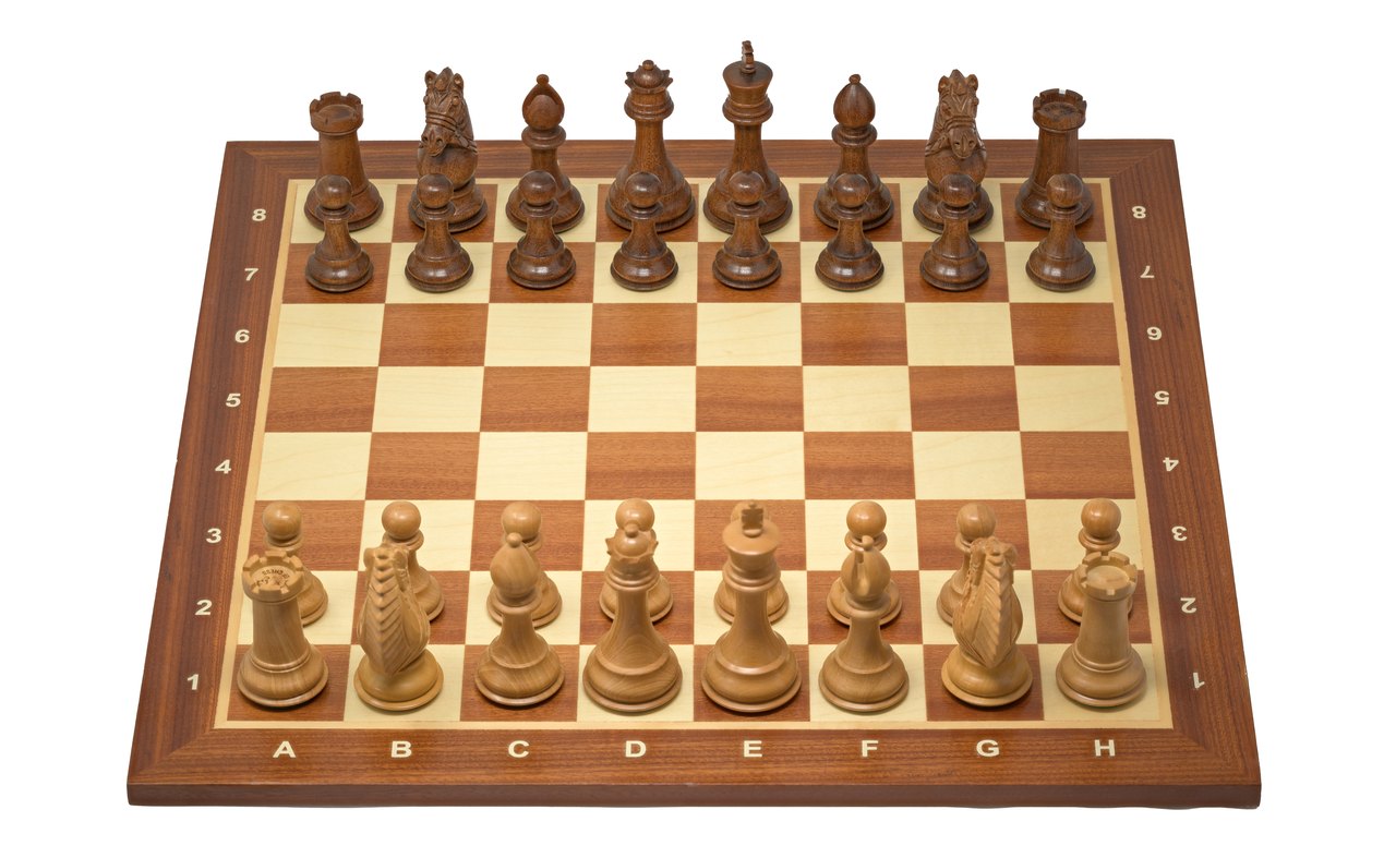 A wooden chessboard photographed from the side at the start of a game, both rows of pieces set out on lettered and numbered squares